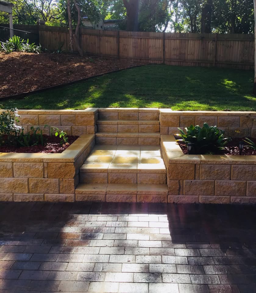 Sandstone-block retaining wall with built-in steps, garden beds either side and brick paving below